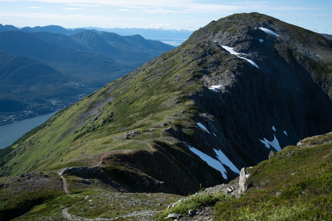 A green, snow-streaked mountain ridge above Juneau, with the city, a channel of water and forested mountains far below.